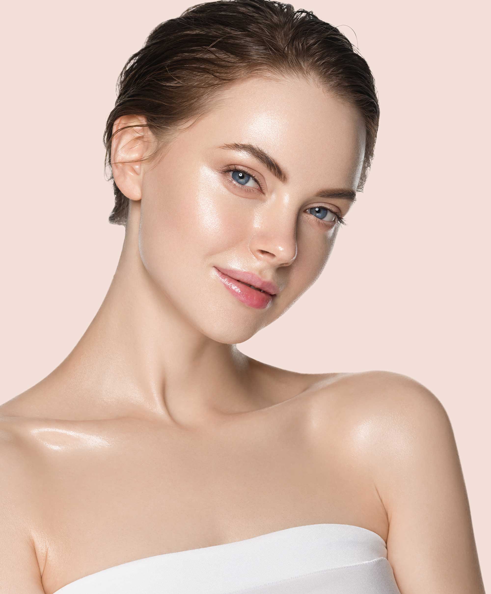 A woman with short, slicked-back hair gazes softly, showcasing radiant skin. She wears a strapless white top against a soft peach background.
